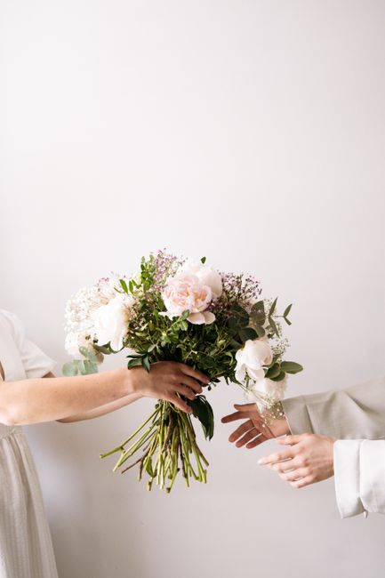 A close-up image of a bouquet of fresh flowers being handed over in a floral gift exchange. The bouquet features large, pale pink roses, white hydrangeas, and smaller purple and white filler flowers,