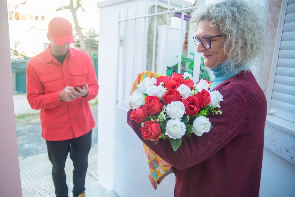 A woman with curly gray hair and glasses holds a vibrant bouquet of fresh red and white roses, wrapped in decorative paper, during a flower delivery. She stands inside near a window with natural light