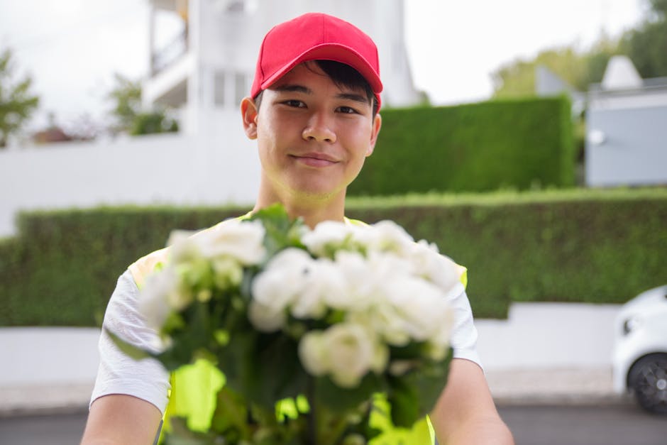 A young male flower delivery courier wearing a red cap and a neon yellow safety vest, smiling as he offers a bouquet of fresh white roses and green foliage. The bouquet is wrapped in clear plastic and
