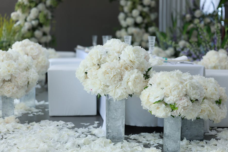 A floral arrangement featuring large, full-bodied white hydrangeas and cream-colored roses displayed in tall, cylindrical, silver metallic vases on a decorated table. The flowers are fresh, with lush