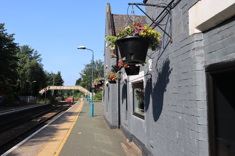 A black hanging flower basket filled with vibrant pink, yellow, and purple petunias and geraniums, suspended from a metal bracket on a grey brick wall of a train station platform, with additional bask