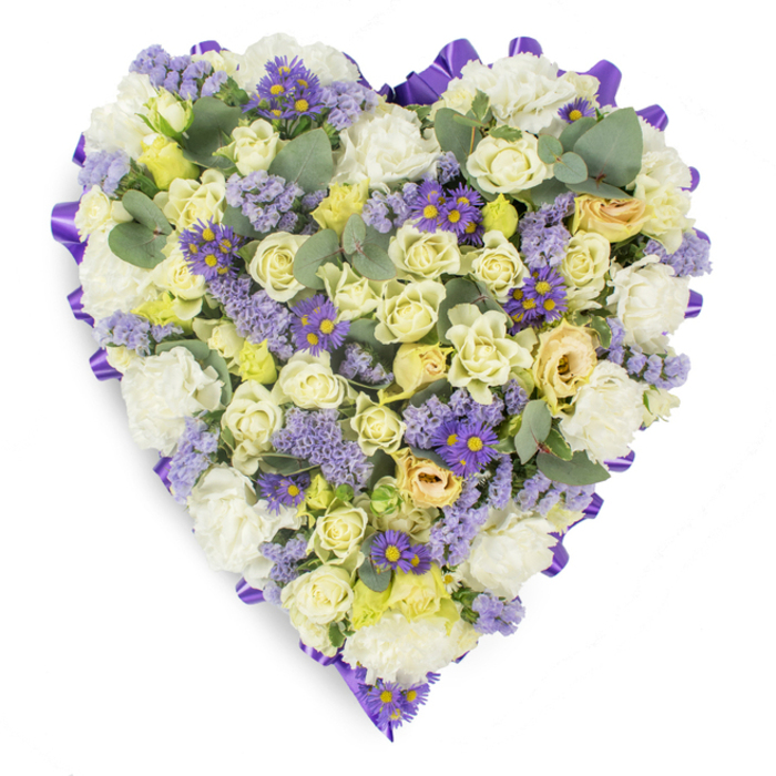 A carefully composed studio image of a heart-shaped floral tribute, presented against a plain white backdrop so the arrangement's form and texture read clearly. A continuous purple satin ribbon loops around the perimeter, its rich tone framing the softer interior like a quiet declaration of care. The interior is a harmonious field of pale, reassuring tones: cream and pale yellow roses dominate, their petals ranging from tight buds to gently unfurling blooms that create a sense of lush softness and subtle movement. Interspersed are generous clusters of white carnations, whose ruffled faces add body and a classic, comforting presence. Delicate lavender statice drifts through the design, providing a light, papery texture and a slightly rustic note, while small purple, daisy-like asters with bright yellow centres offer sharper colour punctuation that livens the composition without diminishing its subdued dignity. Blue-green eucalyptus leaves are tucked strategically among the flowers, their rounded forms and silvery hints breaking the floral mass and suggesting a cool, resinous scent. The even studio lighting highlights the tactile contrasts and the heart's density, reinforcing an atmosphere of peaceful remembrance. This type of memorial heart is commonly chosen for funeral services and graveside gatherings across South Croydon and nearby neighbourhoods such as Selsdon and Purley, where families look for an arrangement that speaks of lasting affection, respect, and consolation. The piece reads as both an exemplar of traditional sympathy floristry and a sensitive, carefully balanced tribute to a loved one.
